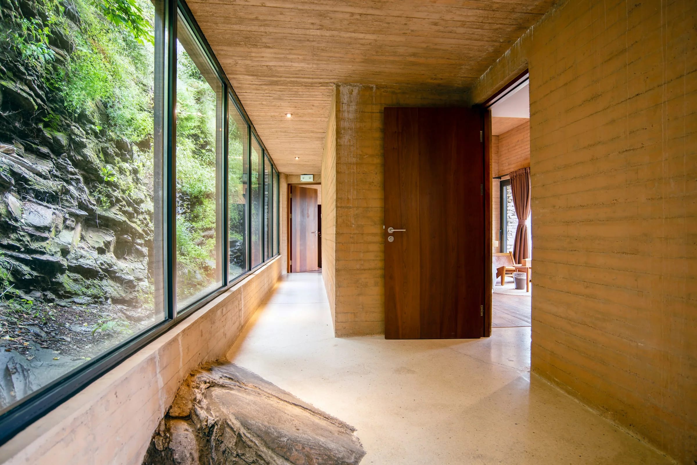 A glazed corridor running tight against a mossy rock face, timber wall on the opposite side
