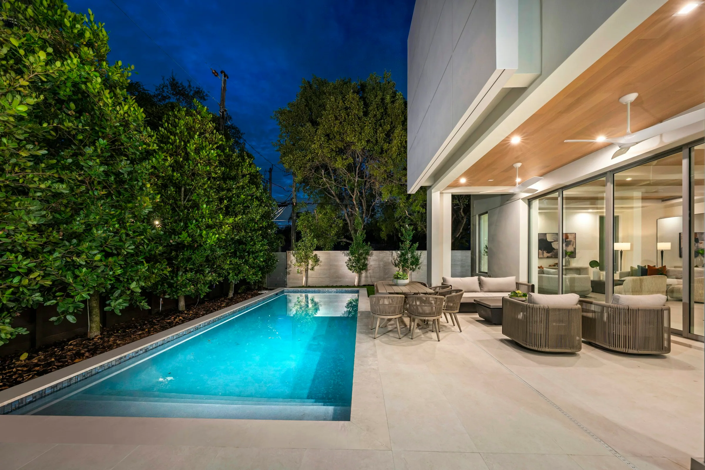 A low modern house at dusk, warm light spilling through full-height glass onto a still pool and an outdoor seating terrace