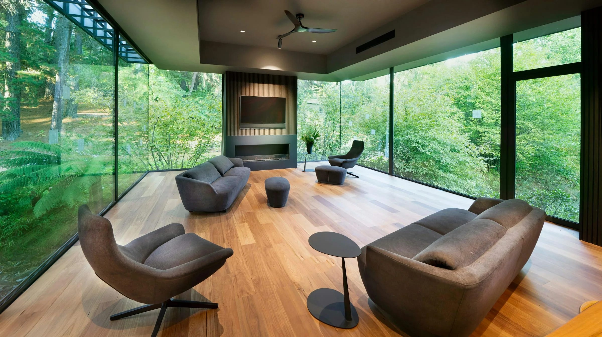A living room with a pale wood floor and glass walls on two sides, looking into dense forest